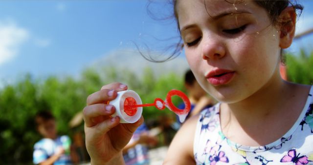 A young girl blowing bubbles in an outdoor summer park with a bright, clear sky. The scene is full of joy and simplicity, capturing the essence of a carefree childhood moment. Ideal for use in advertisements, blog posts about parenting and childhood activities, or promotional materials for outdoor events and playgrounds.