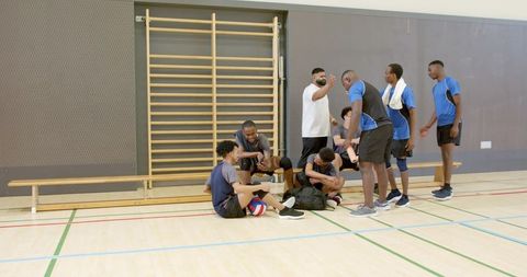 Coach leading diverse male volleyball team during gym training break with bench huddle