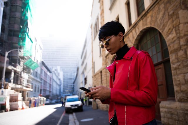 Side view of a fashionable biracial transgender in the street, standing on a street, using smartphone, wearing sunglasses