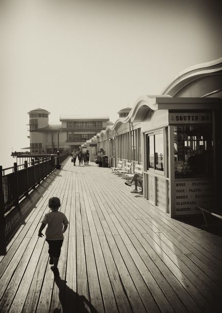 Child running on wooden boardwalk of a pier towards a seaside pavilion. Shops lining boardwalk. Shadow cast by child adds to nostalgic feel. Great for themes of childhood, nostalgia, summer vacations, vintage seaside excursions.