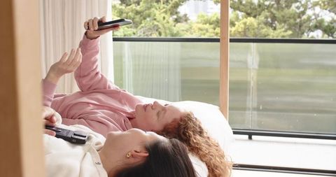Diverse women lying on bed by balcony holding smartphone and tv remote enjoying lazy morning