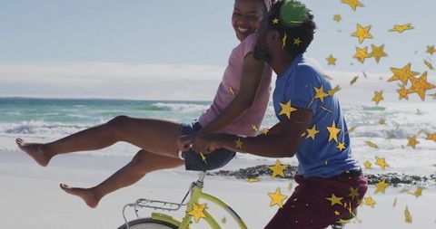Couple riding beach bicycle showered with gold star confetti, playful romantic moment