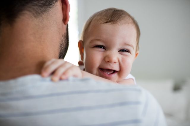 Father holding his smiling baby girl at home, showcasing a tender moment of bonding and happiness. Ideal for use in parenting blogs, family-oriented advertisements, and articles about fatherhood and family life.