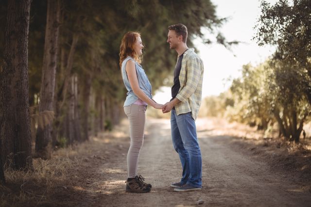 Young couple holding hands while standing on a dirt road at an olive farm. They are facing each other, surrounded by trees, and dressed in casual clothing. This image can be used for themes related to romance, relationships, nature, and rural life.