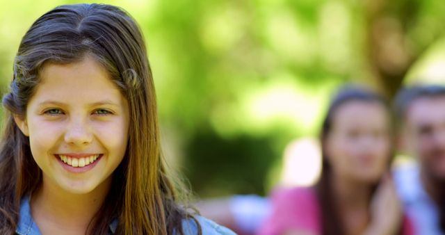 Young girl with long hair smiling outside in a park, with bright green trees and natural sunlight. The background includes blurred figures of people, suggesting family time or community. Ideal for use in ads about outdoor activities, childhood, happiness, family leisure, and summer vacation.