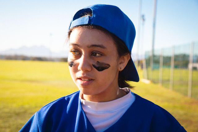 Young female baseball player wearing blue uniform and cap, with black face paint, standing on field during game. Ideal for use in sports-related articles, promotional materials for women's sports, and advertisements for athletic gear.