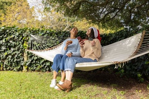 Diverse friends laughing on hammock sharing phone and credit card wearing santa hat