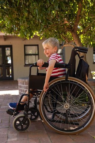 Blonde boy smiling and touching wheelchair rim on sunny brick courtyard with backpack