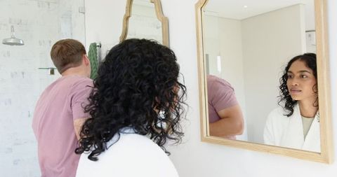 Image shows a diverse couple in a modern, minimalist bathroom. The man is wearing a pink t-shirt, and the woman is in a white bathrobe. The scene highlights mirrors and stylish interiors, which suggests an elegant and tranquil atmosphere. Could be used for topics related to lifestyle, interior design inspiration, and modern home living.