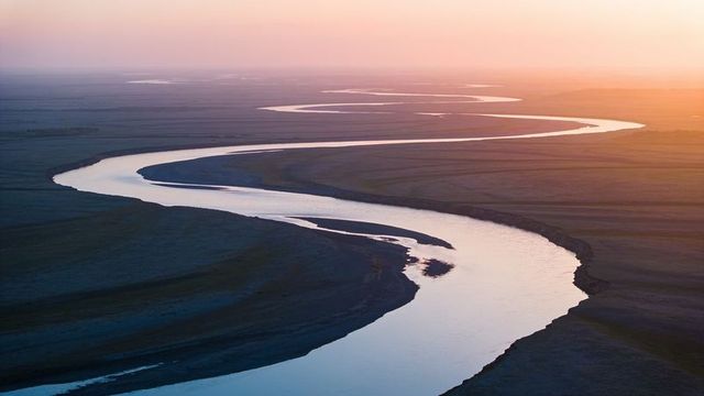 Winding river meandering across vast floodplain at sunrise with soft pastel horizon