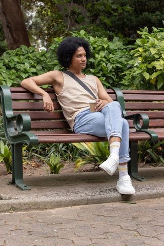 African american man relaxing on park bench holding takeaway coffee cup in green park