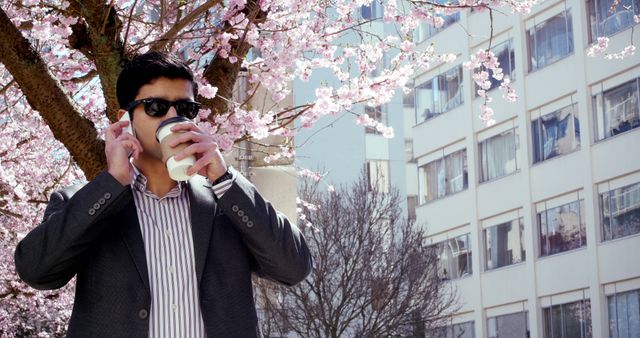 Businessman wearing sunglasses sipping coffee under blooming cherry blossoms on a sunny spring day in an urban area. Can be used for concepts like relaxation, work-life balance, urban nature, city professional enjoying nature, springtime business breaks.