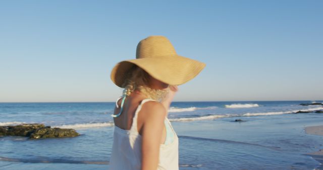 Woman wearing straw sun hat and white summer dress walking along sandy beach with ocean waves beside her. The ocean and clear blue sky create relaxing and sunny seaside environment. Perfect for themes related to summer, travel, leisure, vacation, outdoor activities, and relaxation.
