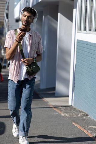 Asian man walking enjoying chocolate muffin listening with headphones on sunlit city sidewalk