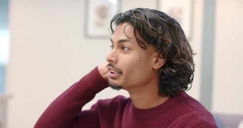 Indian man sitting on sofa in modern lounge wearing maroon sweater touching neck