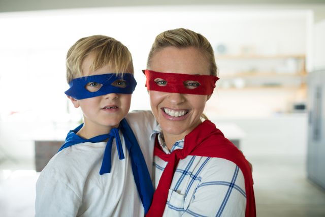 Mother and son dressed as superheroes with masks and capes, enjoying playful bonding time in living room. Ideal for themes of family fun, childhood imagination, and parent-child relationships. Suitable for use in parenting blogs, family-oriented advertisements, and educational materials.