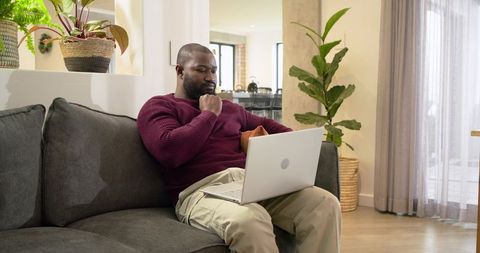 African American man working from home on laptop on gray sofa in modern open-plan living room