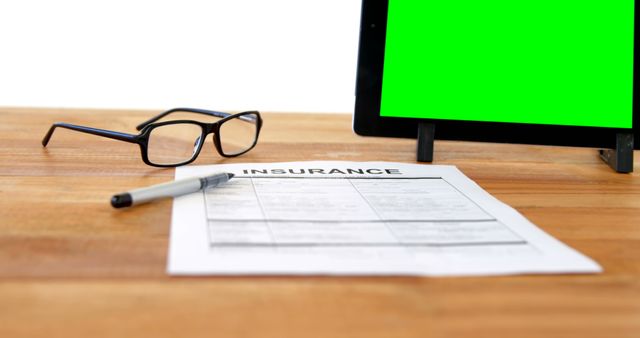 A pair of glasses and a pen rest beside an insurance form on a desk, with a tablet displaying a green screen in the background, with copy space. It suggests a professional setting, for a business or insurance consultation.