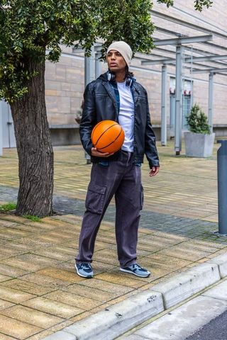 Young man holding basketball on urban plaza wearing leather jacket and headphones