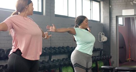 Diverse women stretching together in gym near dumbbells wearing mint green and pink tops