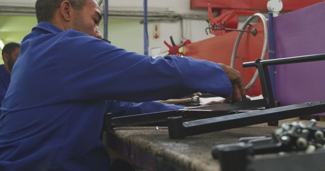 Factory worker is meticulously assembling mechanical parts at a workbench in an industrial workshop. The worker's blue uniform signifies a professional setting in manufacturing or heavy industry. Viable for articles on industrial processes, manufacturing, labor, and craftsmanship. Useful for illustrating topics on precision engineering, industrial work environments, and assembly line jobs.