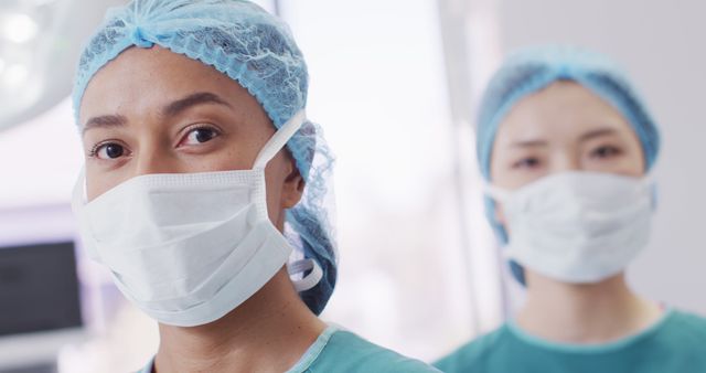 Two healthcare professionals are standing in a hospital environment, wearing surgical masks, hairnets, and green scrubs. This picture can be used for articles related to health and safety in medical settings, promotional materials for hospitals or healthcare facilities, or educational content about medical protocols and the importance of protective gear in healthcare.