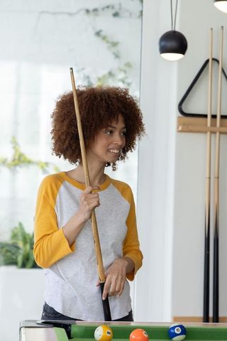 African american woman holding pool cue and smiling in contemporary home game room with mustard top