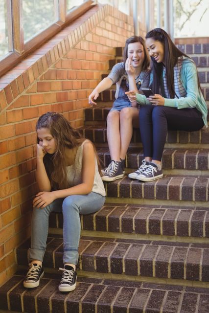 Teenage girls sitting on school staircase, with two girls laughing and one girl looking sad and excluded. Useful for illustrating themes of bullying, peer pressure, social exclusion, and mental health in educational settings. Can be used in articles, blogs, and campaigns addressing school bullying and its impact on teenagers.