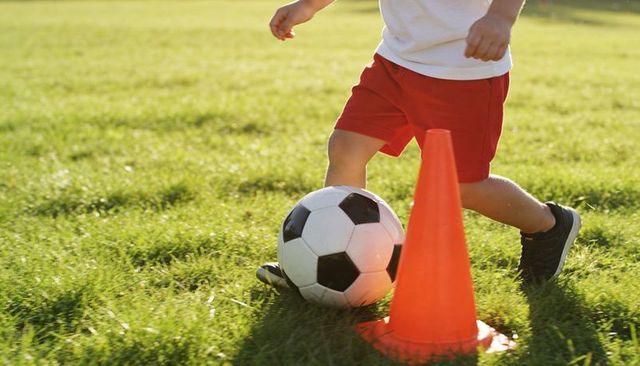 Dribbling youth practicing soccer drills on sunlit grass with ball and orange cone