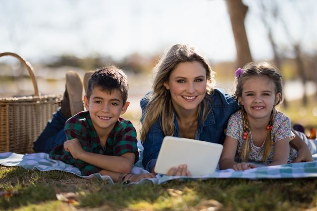 Mother and two children lying on blanket in park, using digital tablet. Ideal for family, technology, and outdoor lifestyle themes. Perfect for advertisements, blogs, and articles about family bonding, digital learning, and leisure activities.