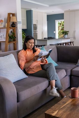 Asian woman reading paperback on gray sectional sofa in modern cozy living room