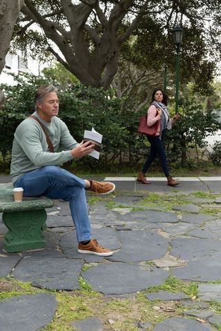 Mature man reading on bench while woman walks by using smartphone in leafy park
