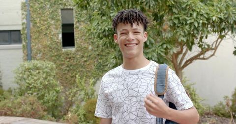 Smiling teenage student standing in campus courtyard carrying denim backpack wearing patterned tee