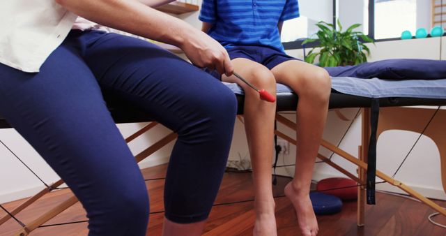 Physiotherapist using a reflex hammer to check the knee reflexes of a child sitting on a padded therapy table. Ideal for illustrating healthcare services, pediatric physiotherapy, and medical examinations in clinics. Can be used in health and wellness websites, educational materials, and informational content on child health checkups.