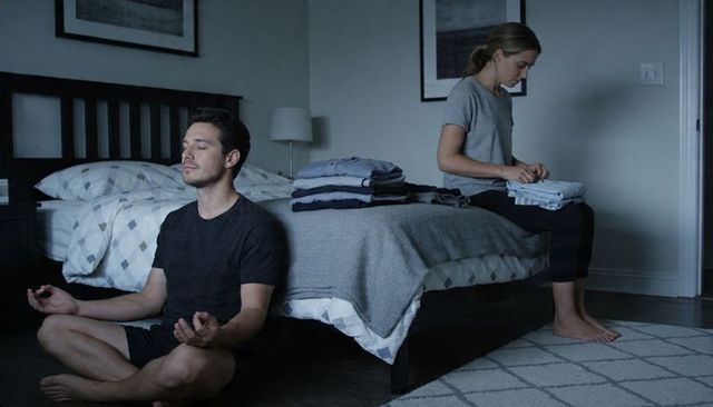Calm domestic ritual: man meditating while woman folding laundry on bed in minimal bedroom