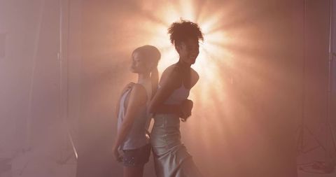 Backlit Fashion Duo Posing Back-to-Back in Hazy Amber Glow with Satin and Sparkle