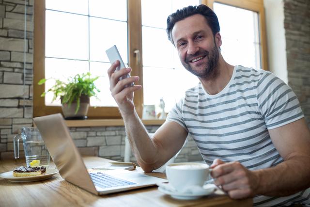 Man smiling while holding a mobile phone and a coffee cup, sitting in a coffee shop with a laptop on the table. Ideal for use in advertisements for coffee shops, technology products, remote work, or lifestyle blogs.