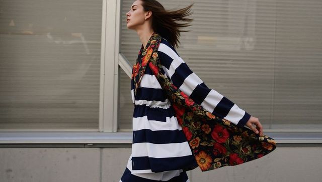 Striding Model Wearing Navy Stripes and Bold Floral Coat Along Urban Glass Facade in Motion