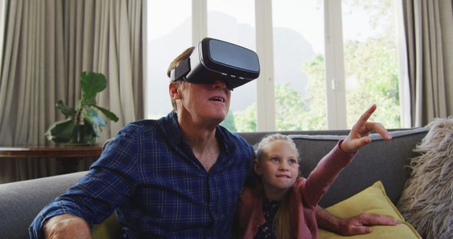 Grandfather sharing a moment with his young granddaughter while using a VR headset. Perfect for depicting multi-generational technology use, family bonding, and modern entertainment at home. Ideal for articles on virtual reality, family dynamics, and tech-savvy seniors.