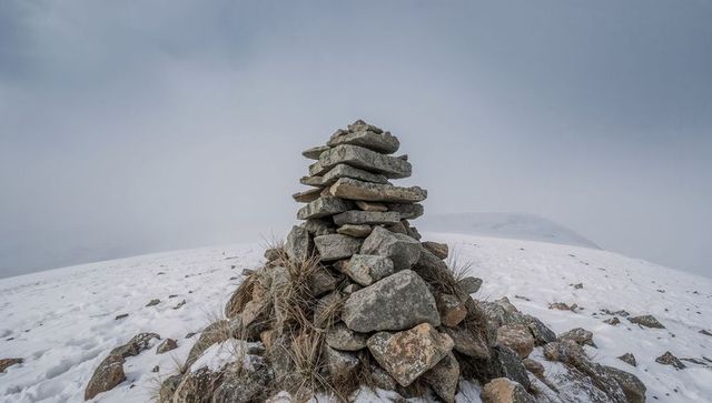 Stacked stone cairn standing on snowy windswept mountain ridge in misty alpine plateau