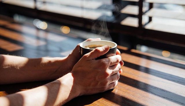Hands cradling steaming mug on sunlit wooden table with blinds and shadows