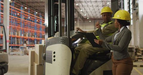 Warehouse supervisor directing forklift operator with clipboard and safety helmets