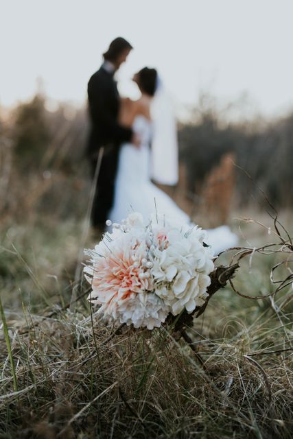 Bride and groom embracing outdoors in a scenic, nature location with blurred faces, bringing attention to a flower bouquet in the foreground. Suitable for wedding invitations, romantic announcements, greeting cards, wedding blogs, or websites related to wedding planning and photography.