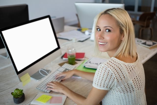 Young woman smiling while working at a computer in a creative office environment. Ideal for use in business, technology, and workplace productivity contexts. Suitable for illustrating modern office settings, professional work environments, and employee engagement.