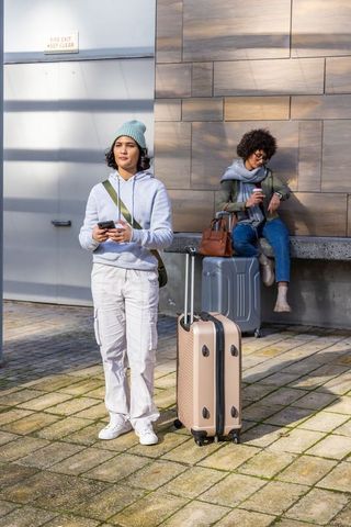 Young women waiting with carry-on suitcases and smartphones in sunlit urban plaza