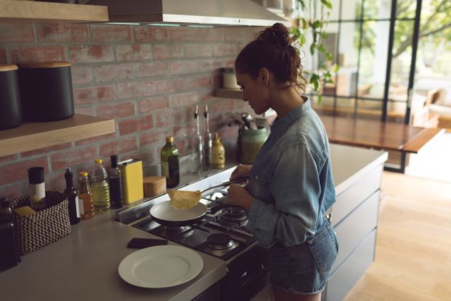 Woman standing at stove cooking in a modern kitchen with exposed brick wall. Suitable for depicting domestic life, home cooking, kitchen design, and casual lifestyle concepts.