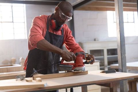Mature african american craftsman sanding plank in workshop wearing goggles and earmuffs
