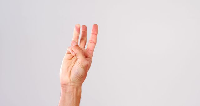 A Caucasian hand is shown making a three-finger gesture against a plain background, with copy space. Such a gesture can signify various meanings, from the number three to a symbol of peace or victory in different cultures.