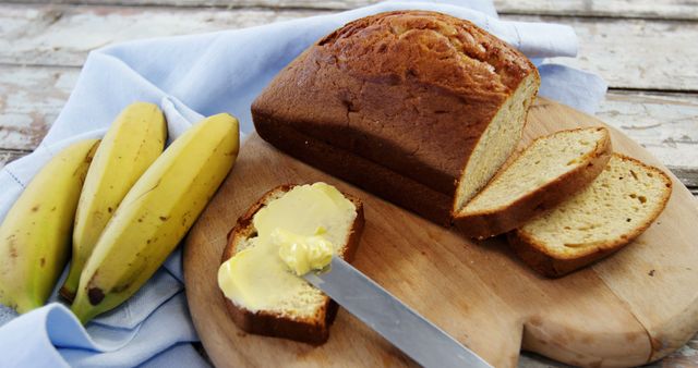A freshly baked loaf of bread is sliced on a wooden board, with a spread of butter on one piece and bananas on the side. Capturing a moment of simple culinary delight, the image evokes the warmth of homemade baking and the appeal of a comforting snack.