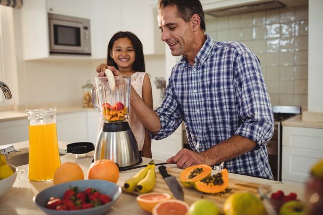 Father and daughter preparing a smoothie together in a modern kitchen. The father is operating the blender while the daughter watches with a smile. Various fresh fruits like bananas, strawberries, and papaya are spread out on the counter. This image is perfect for illustrating family bonding, healthy eating, and home cooking. It can be used in articles about parenting, nutrition, or lifestyle blogs.